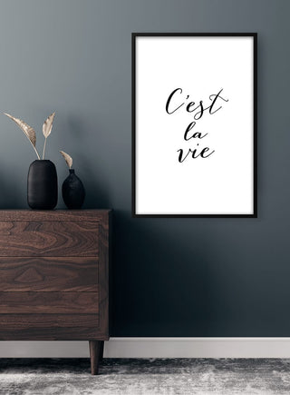 C'est la Vie poster in black script on a white background, framed and displayed above a modern wooden dresser.