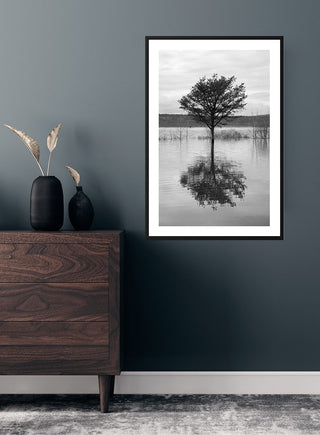 Black and white poster of a tree reflected in a lake, framed and hanging above a wooden dresser.