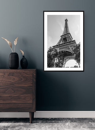 Black and white poster of the Eiffel Tower viewed from below, framed and hung on a dark wall above a wooden dresser.