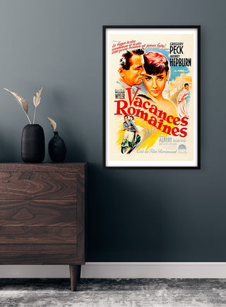 A framed "Vacances Romaines" Audrey Hepburn movie poster displayed on a dark wall above a wooden dresser.