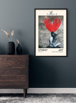 Framed Banksy-inspired street art print with a red heart displayed on a gray wall above a wooden dresser.