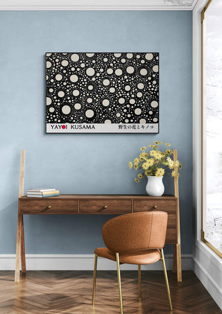 Framed Yayoi Kusama - Black Dots Poster hangs above a wooden desk with a vase of yellow flowers in a modern room.