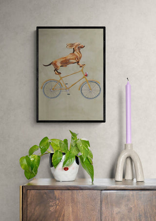 Framed artwork of a dachshund riding a bicycle, displayed above a modern cabinet with decor.